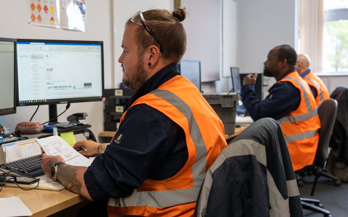 Man in high-vis jacket holds paper while working at a computer