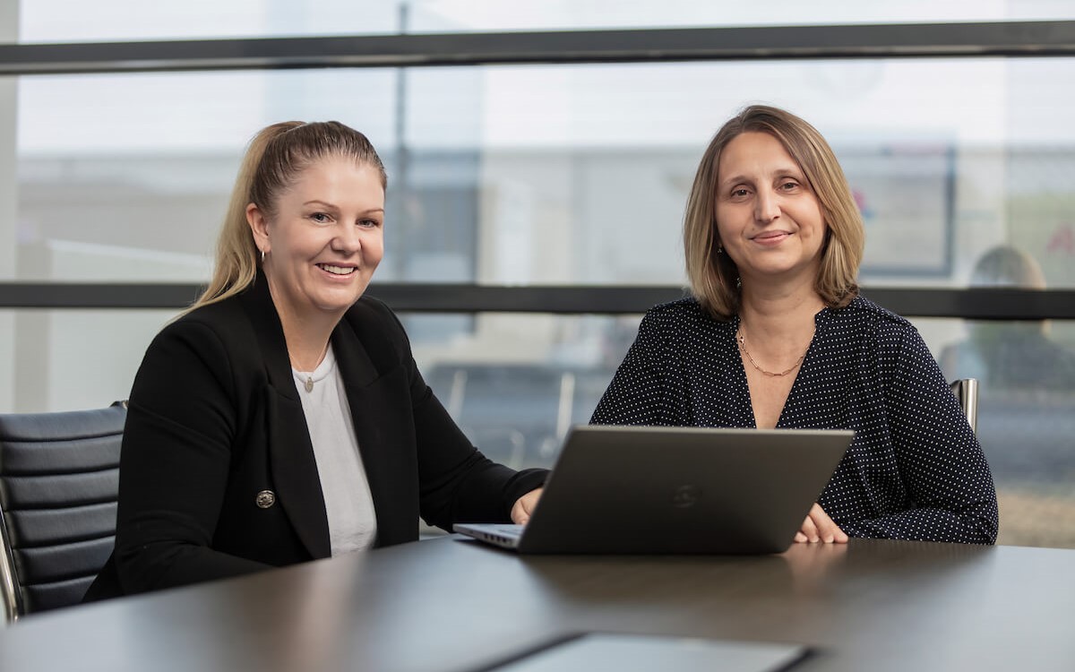 Two women sat by a laptop smiling at the camera