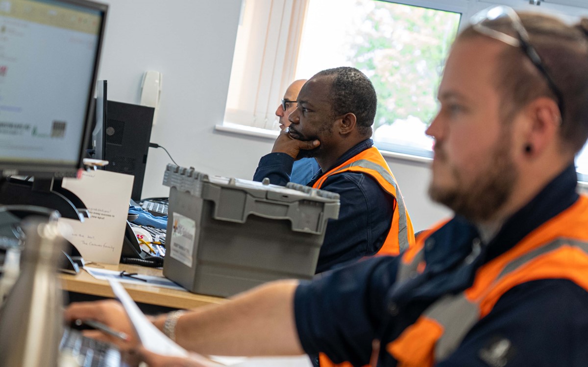 Three men in orange high-vis jackets work at computers in an office