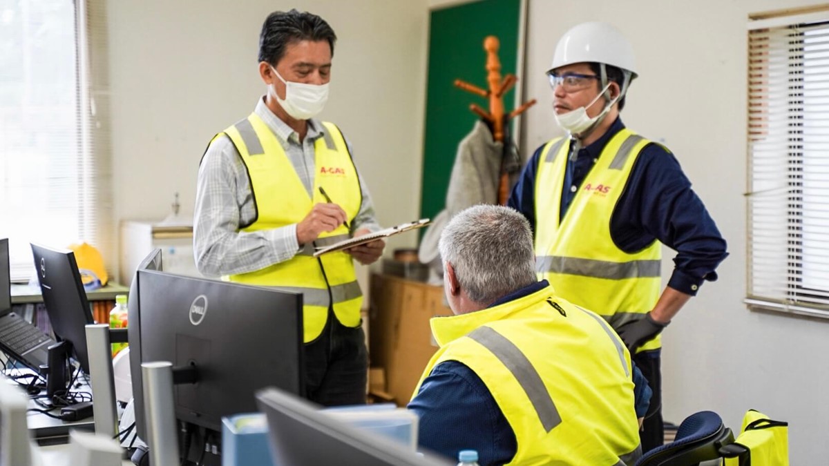Three men in yellow high-vis jackets talking in and office