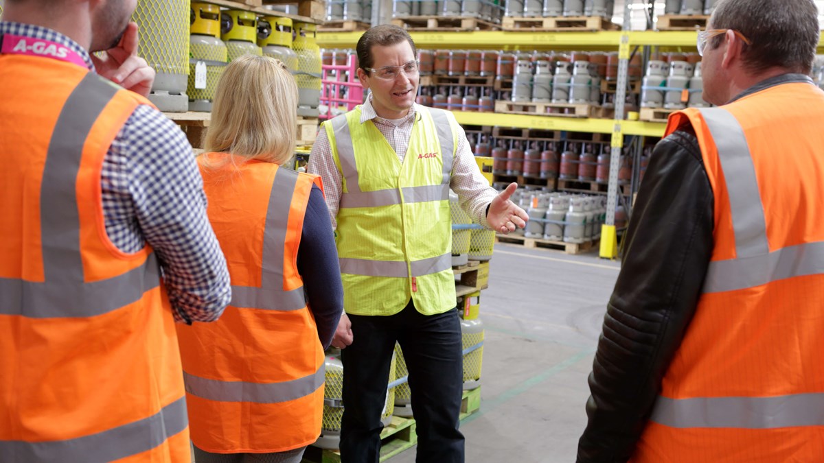 A man in a hi vis giving a site tour to visitors, we can only see the back of visitors heads, the man is surrounded by palettes of gas cylinders