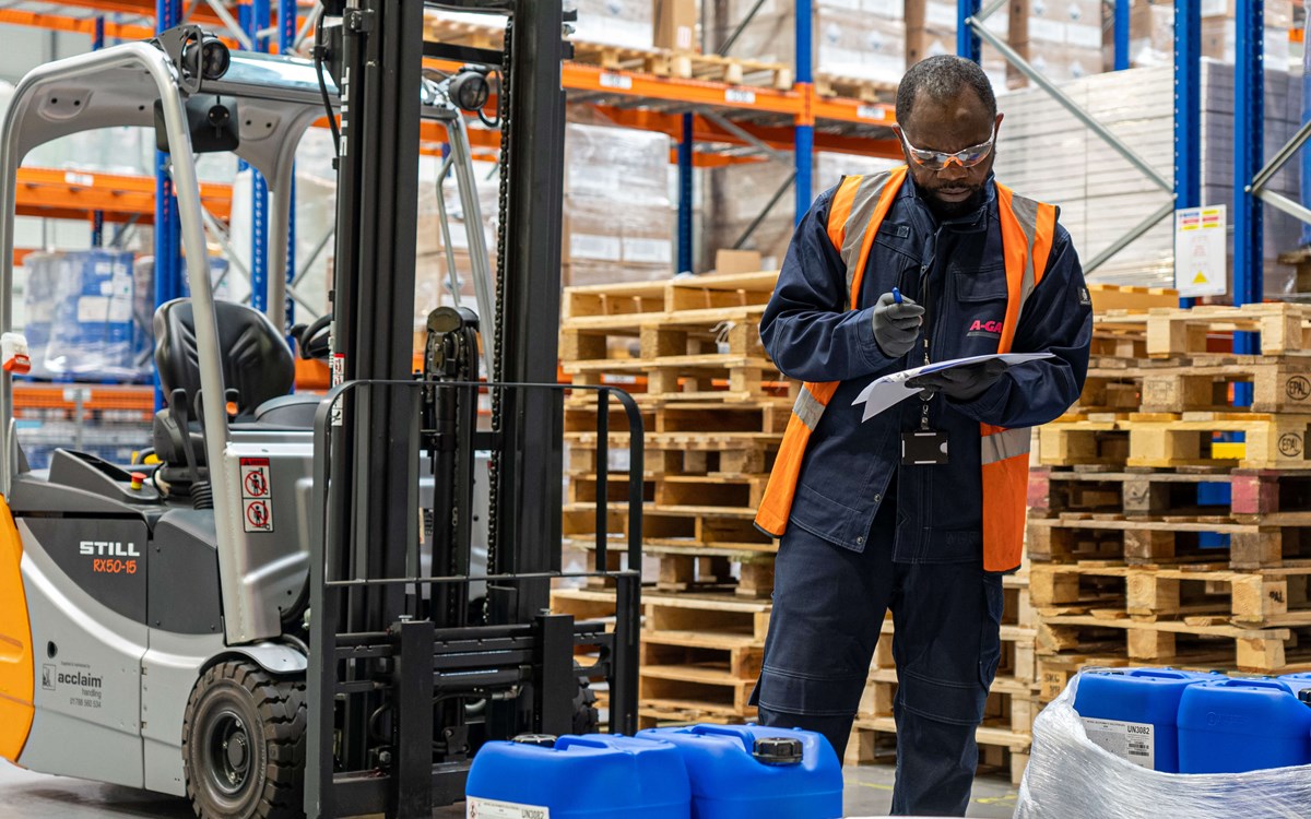 A man in a high-vis orange jacket writing on a clipboard in front of a forklift truck in warehouse